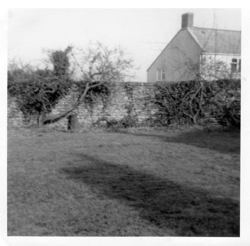 An old black and white photograph of a lawn area with a wall behind and part of a farmhouse. The field is empty of people.