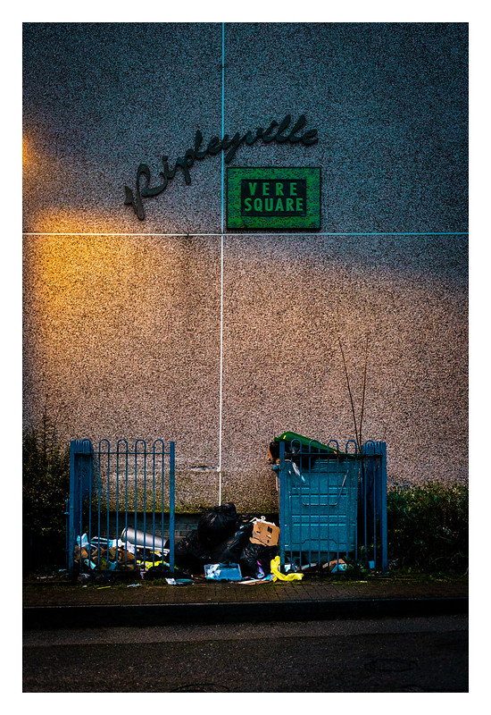 A picture of a sign for a housing estate saying Ripleyville Vere Square in ornate writing. Underneath is an overflowing refuse bin