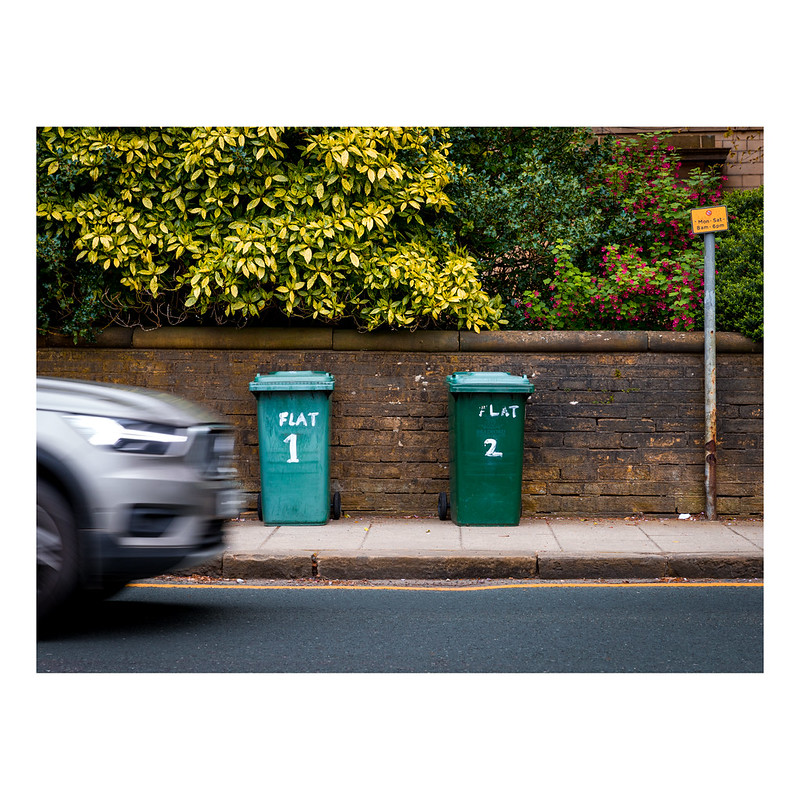 Two wheelie Bins labelled Flat 1 and Flat 2 in front of a low wall made of Yorkshire Stone. The garden shrubbery fills the top of the picture. In the front of the picture is the front of a grey car which is slightly blurred due to its speed.