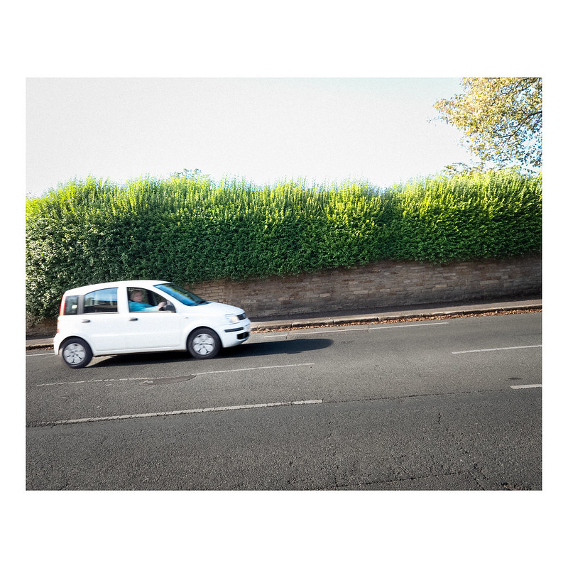 A side on view of a white car driving up a hill. Behind is a low stone wall and a very bushy hedge that is lit up at the top by the sun. The car driver is just about visible but blurred, it is a woman looking suspiciously at the photographer.
