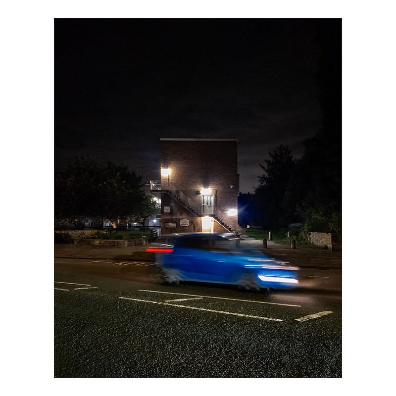 A night time photograph showing a side view of a blue car with headlights on that is blurred because of the movement. Behind is a modern low rise residential block with steps and lights visible.