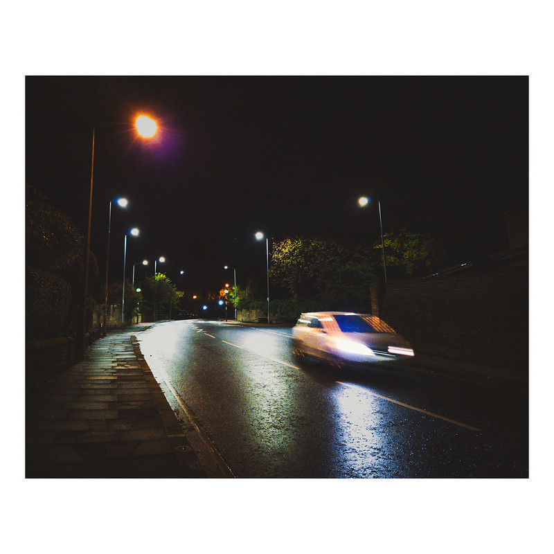A night time photograph showing a blurred white car driving downhill. The road is wet and there are reflections from streetlights. Just about visible are hedges in front of the houses each side of Park Road as we look uphill.