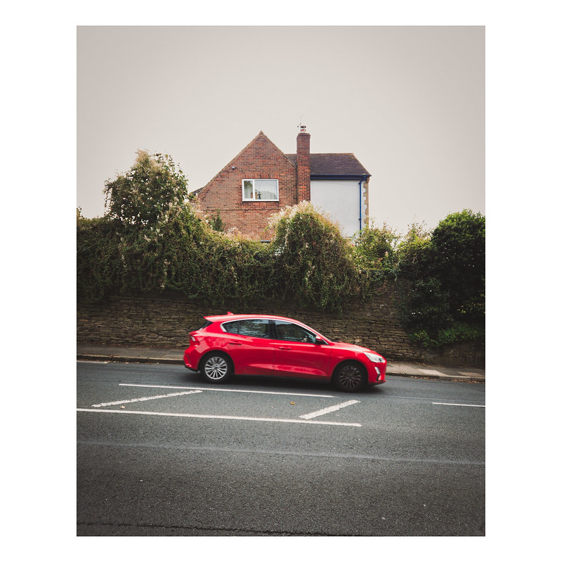 A red car driving on a road downhill. Behind is a wall with a hedge on top that is very uneven. Behind the hedge, the top of a house can be seen.