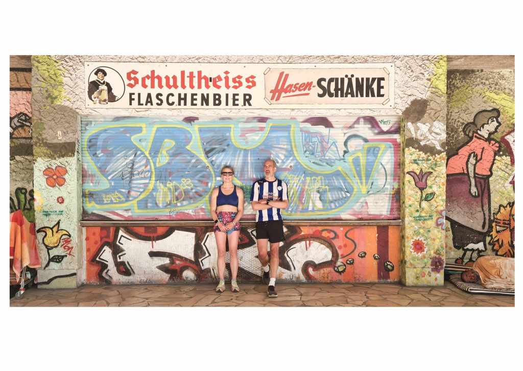 Two people wearing shorts and sportswear are posing looking very cool in front of a cafe bar that is shuttered. The shutter and concrete structure of the cafe is covered in graffiti. At the right and left of the picture it is possible to make out homeless people sleeping