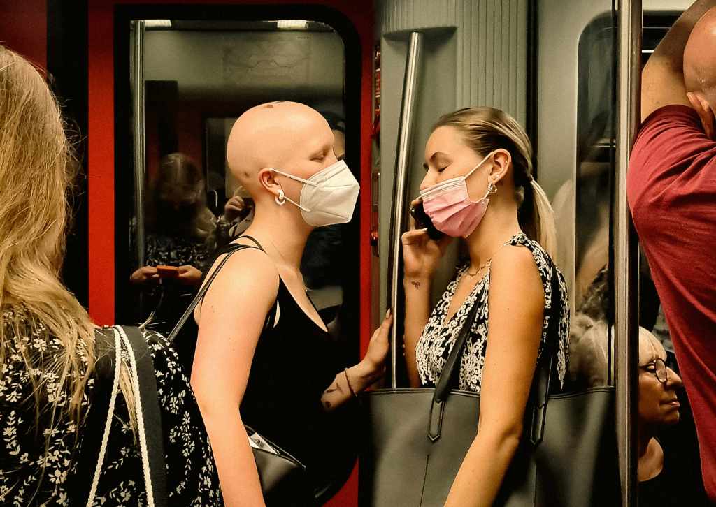 A single picture taken in an underground railway carriage (U-Bahn). The train is crowded and the main subject is two women who are standing close to each other, as if they are travelling together. One woman, who is bald is looking lovingly at the other, who is speaking on her phone and not looking back at her companion. Both women look tired. Both women and some of the other passengers are wearing masks,