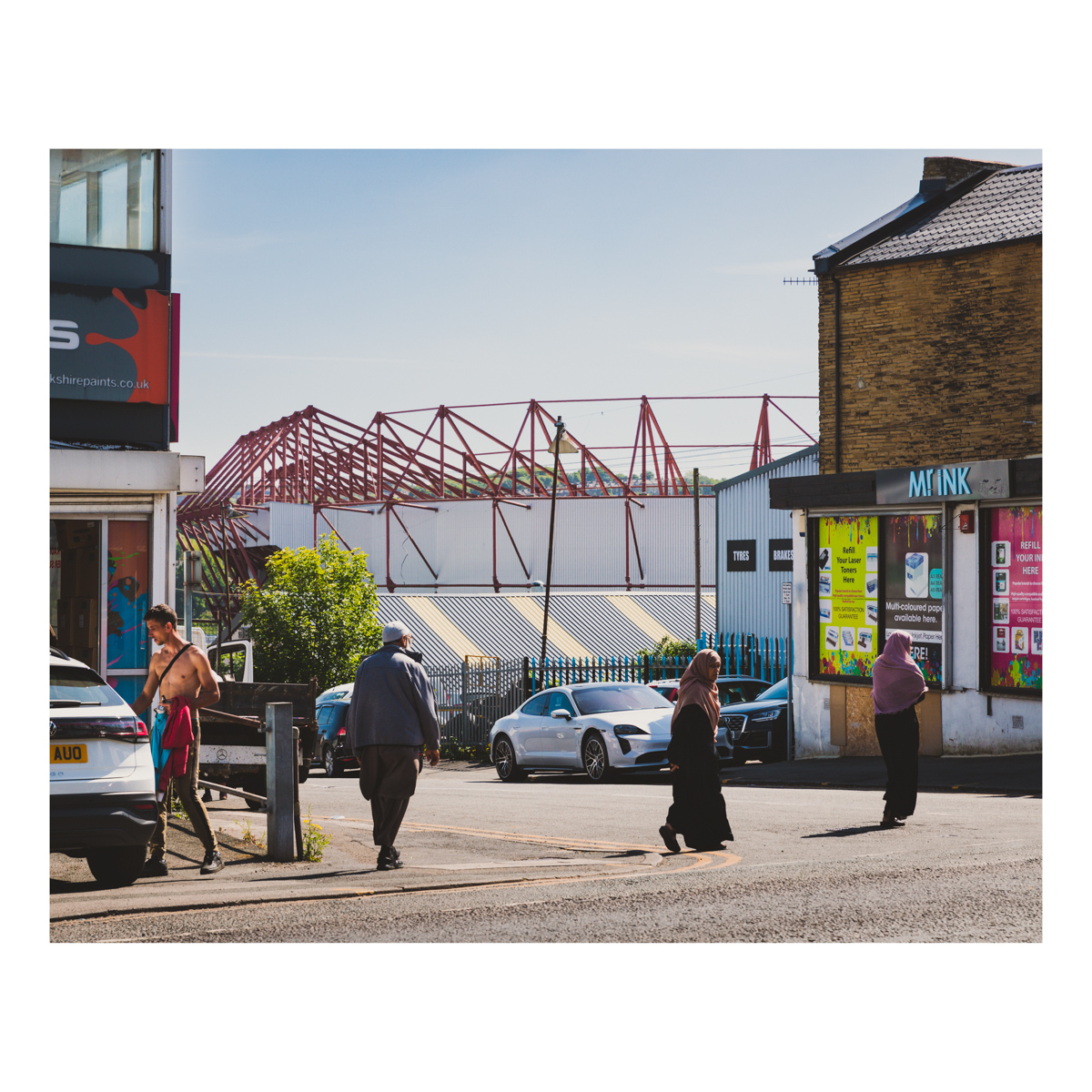 A view of the Bradford City football stadium from Manningham Lane. Crossing a side road in front of a shop named Mister Ink are two women dressed in Hijab and a gentleman in a naweeb and kufi hat. Behind the man there is a topless white man getting into a car.